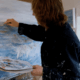 A person with shoulder-length hair wearing a dark, paint-splattered shirt paints an abstract seascape on a canvas. They hold a palette with mixed colors and stand in a bright art studio. Sancreed House Retreat