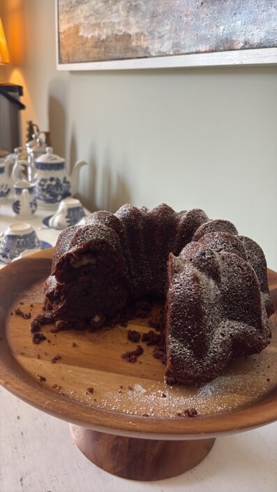 A sliced chocolate Bundt cake dusted with powdered sugar sits on a wooden cake stand, with teapots and cups in the background on a white table. Sancreed House Retreat