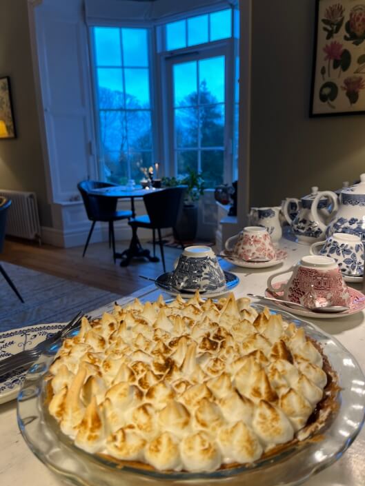 A pie with toasted meringue sits on a table set with patterned teacups and teapots. In the background, a dining table is set near large windows overlooking trees at dusk. Sancreed House Retreat
