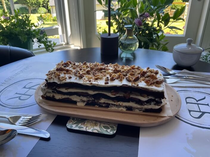 A layered dessert with cream and crumbled topping sits on a wooden platter atop a table by a window, surrounded by cutlery, placemats, a sugar bowl, and green plants outside. Sancreed House Retreat