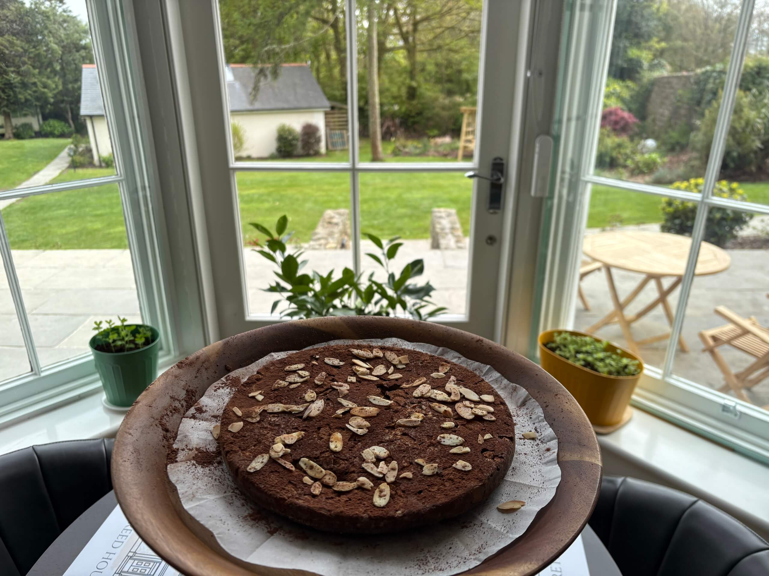A chocolate cake topped with oat flakes sits on a wooden plate by a large window, with potted plants on the windowsill and a view of a green garden and patio furniture outside. Sancreed House Retreat