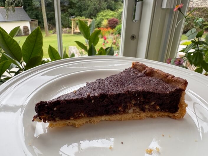 A slice of chocolate tart with a golden crust sits on a white plate. Behind it, a window shows a lush green garden with trees, bushes, and a lawn. Bright natural light illuminates the scene. Sancreed House Retreat