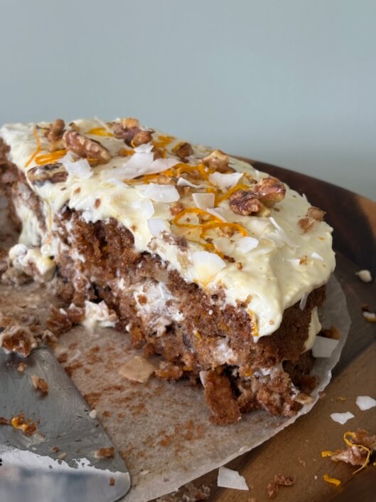 A partially sliced carrot cake with cream cheese frosting, topped with walnut pieces, coconut flakes, and orange zest, sits on parchment paper on a wooden surface. A cake server rests nearby. Sancreed House Retreat