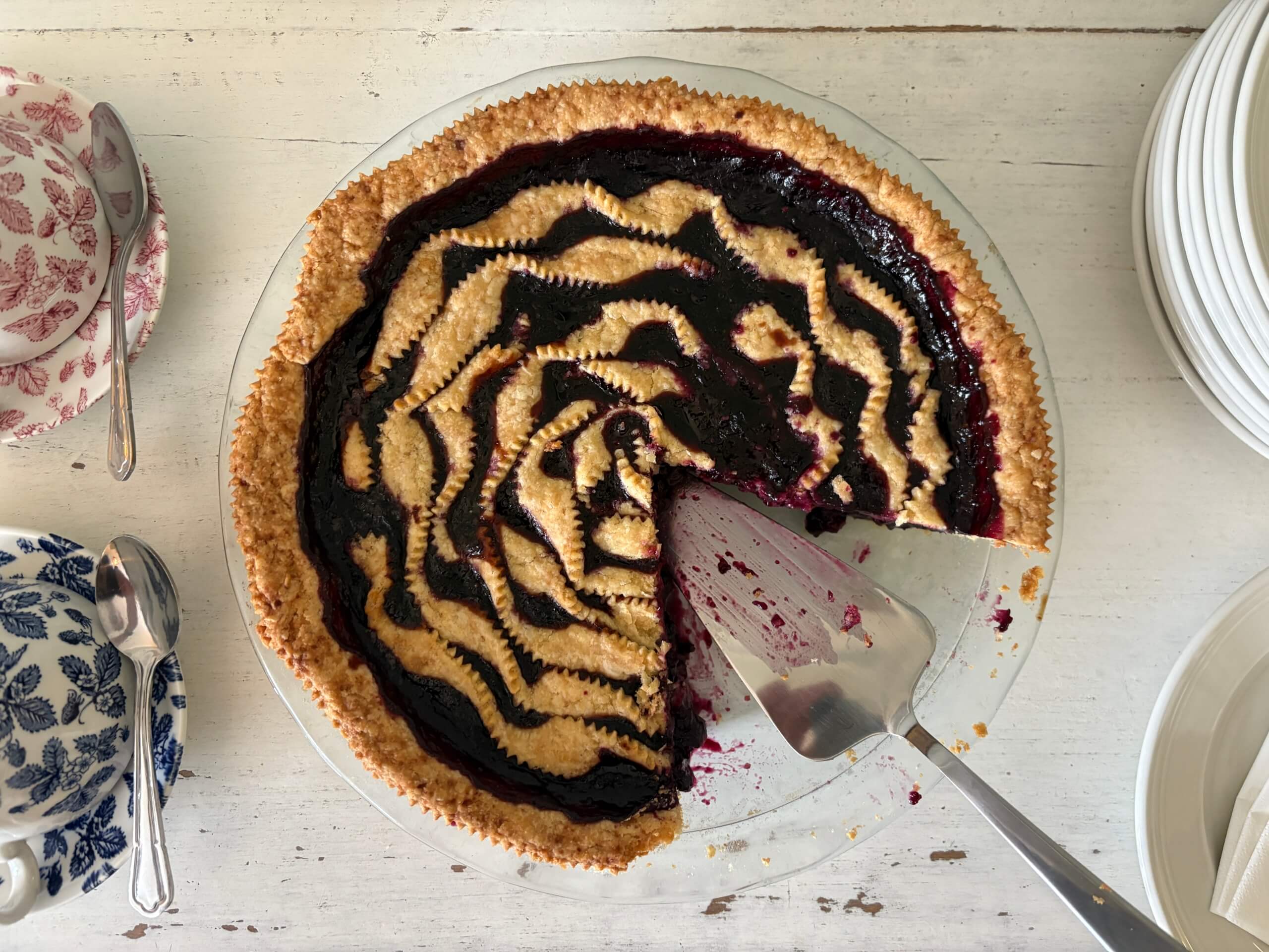 A round pie with a golden, wavy lattice crust and dark berry filling sits on a white table. A slice has been removed with a pie server. Nearby are stacked plates and two floral teacups with matching saucers and spoons. Sancreed House Retreat