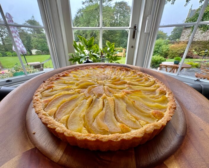 A freshly baked pear tart sits on a wooden platter in front of a large window, with a garden and patio visible outside on a cloudy day. Sancreed House Retreat