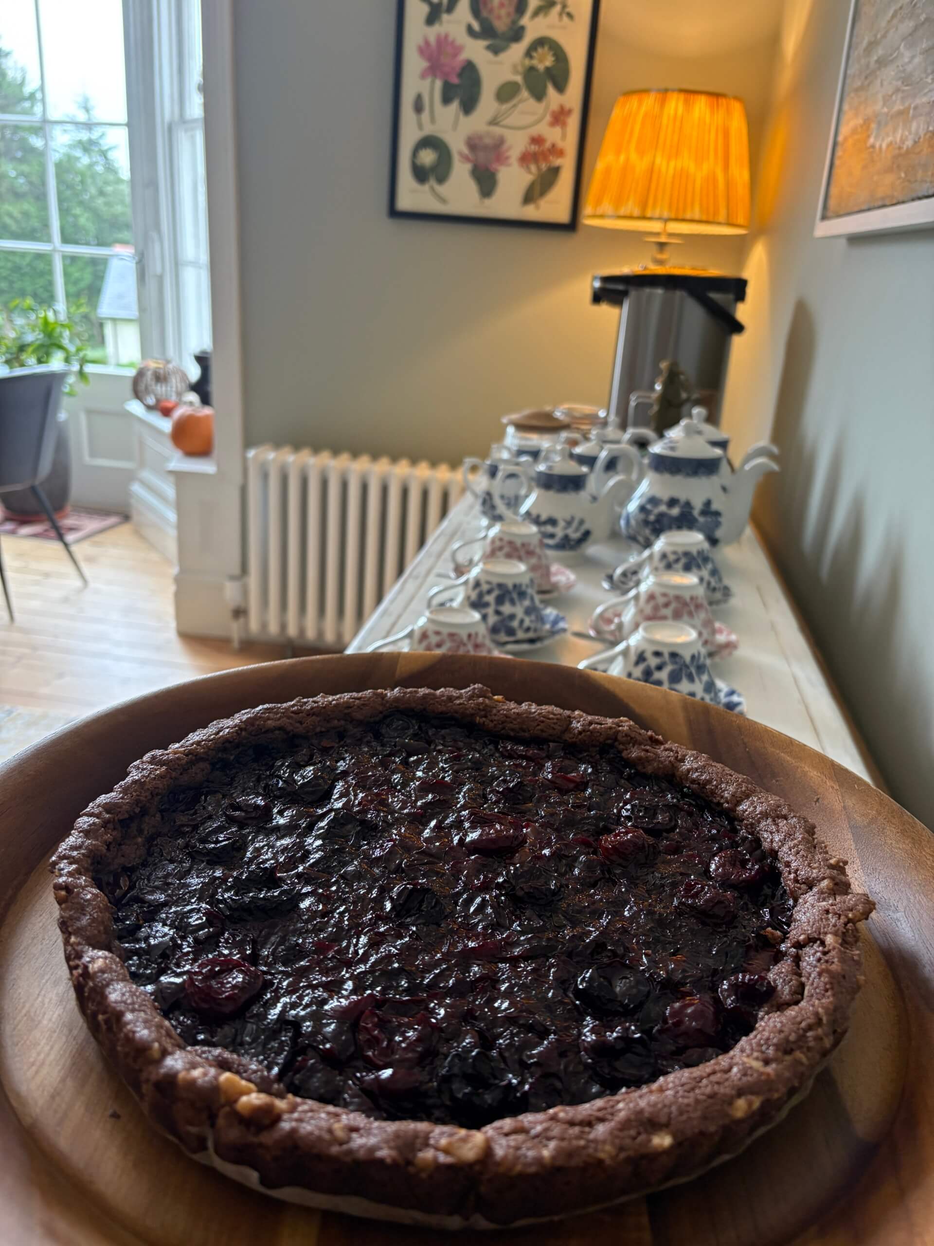 A chocolate berry tart on a wooden plate sits in the foreground. In the background, a tea set, teapots, and cups are arranged on a sideboard beneath a lit lamp in a cozy, softly lit room. Sancreed House Retreat