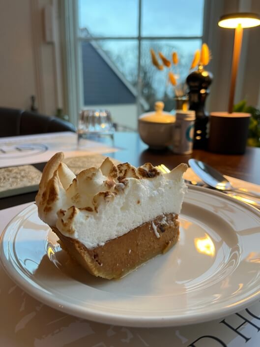 A slice of pie with a golden-brown meringue topping and pumpkin filling sits on a white plate, on a dining table near a window with soft daylight and tableware in the background. Sancreed House Retreat