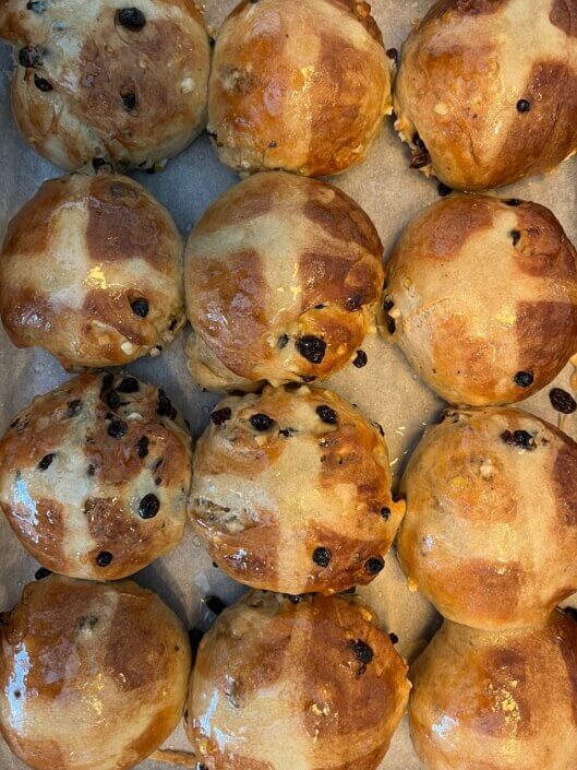 A close-up view of freshly baked hot cross buns arranged in rows, featuring shiny golden-brown tops with crosses and dotted with currants or raisins. Sancreed House Retreat