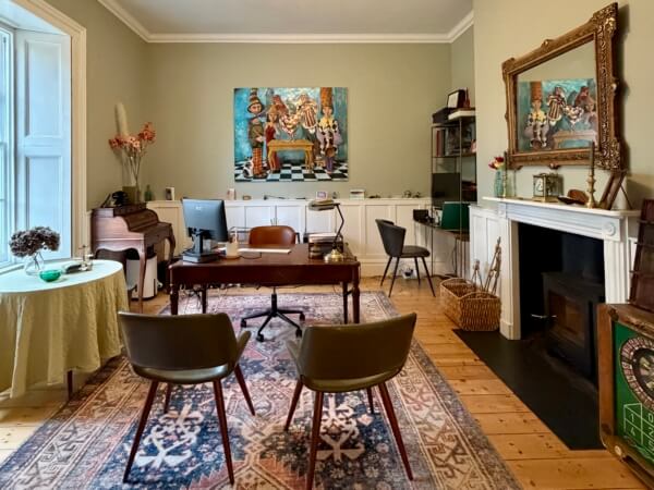 A cozy home office with a large rug, two chairs facing a wooden desk, a fireplace with a ornate mirror, a piano, and a colorful painting featuring animals in a whimsical scene on the back wall. Sancreed House Retreat