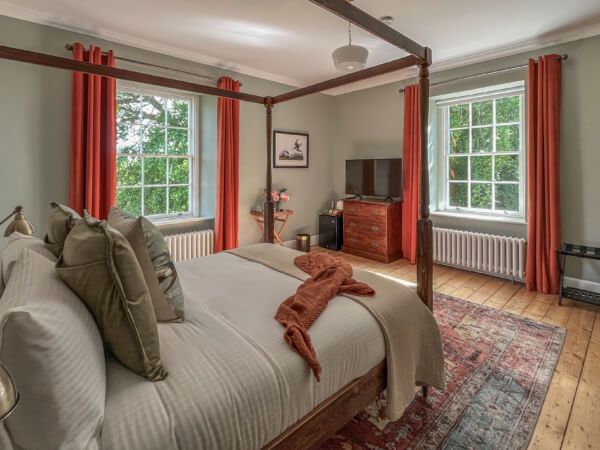 A bright bedroom with a four-poster bed, white bedding, orange pillows, and throw. Two large windows with orange curtains, a dresser with a TV, and a patterned rug are visible. The room has wooden floors and green walls. Sancreed House Retreat