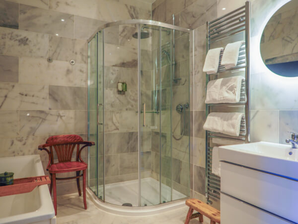 Modern bathroom with marble walls, a glass-enclosed shower, a towel rack with folded white towels, a white sink, a round mirror with lighting, a wooden chair, and a small wooden stool. Sancreed House Retreat