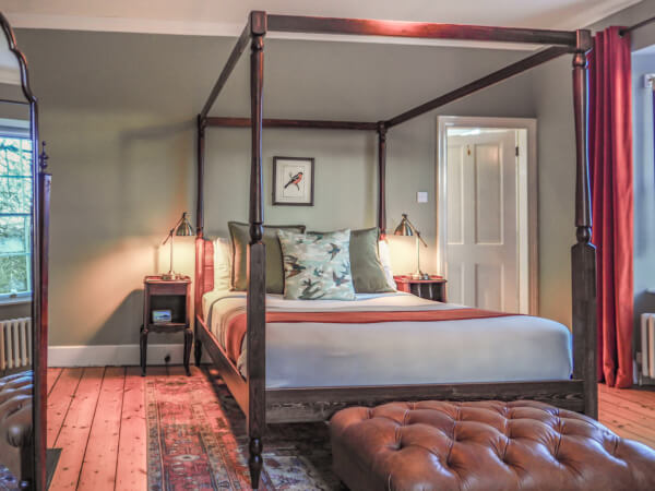 A cozy bedroom with a dark wooden four-poster bed, patterned cushions, bedside tables with lamps, a large window, a tufted leather ottoman, and a colorful rug on a wooden floor. Sancreed House Retreat