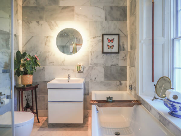 A modern bathroom with marble walls, a round lit mirror above a white sink, a bathtub with a wooden tray, a potted plant on a small table, and framed butterfly art on the wall. Natural light streams in from a window. Sancreed House Retreat