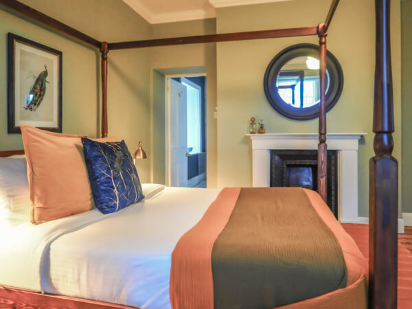 A cozy bedroom with a four-poster bed featuring white linens, brown and blue pillows, and a brown throw. The room has green walls, a round mirror above a white fireplace, and a framed artwork. Sancreed House Retreat