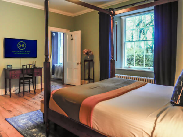 A cozy bedroom with a four-poster bed, green walls, a desk with a chair, a TV on the wall, and large windows letting in natural light with a view of trees outside. Sancreed House Retreat