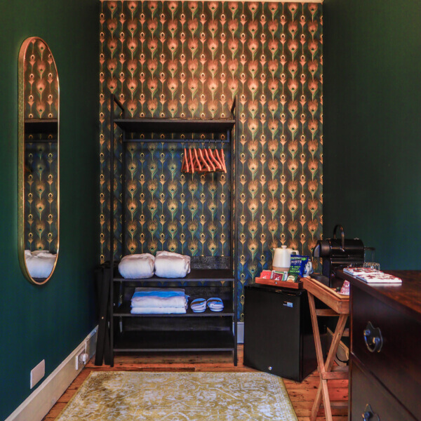 A cozy room with green patterned wallpaper, an open wardrobe with shelves holding folded towels, a mirror on the left wall, a mini fridge, a coffee machine, and a wooden tray table with snacks and drinks. Sancreed House Retreat