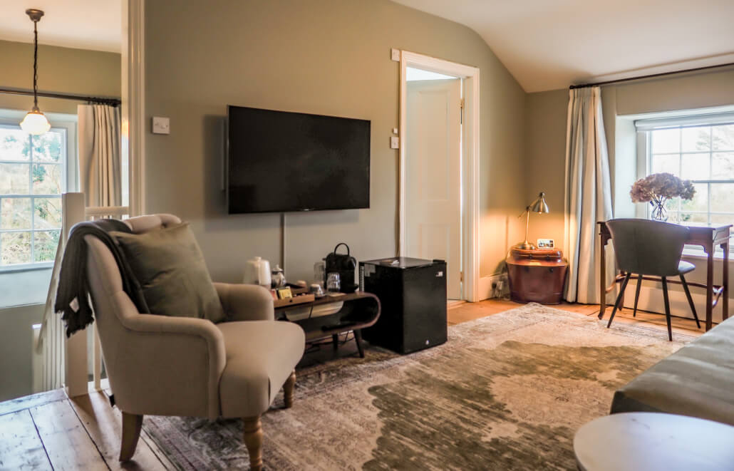 A cozy living room with a beige armchair, wall-mounted TV, small black fridge, table with a lamp and flowers, large rug, and sunlight coming through windows with light curtains. Sancreed House Retreat