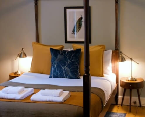 A cozy bedroom with a neatly made four-poster bed, mustard yellow and blue pillows, folded towels, bedside tables with lamps, an armchair, and framed bird artwork on the wall. Sancreed House Retreat